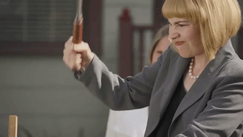 A blond woman in a blazer hammers a piece of wood awkwardly