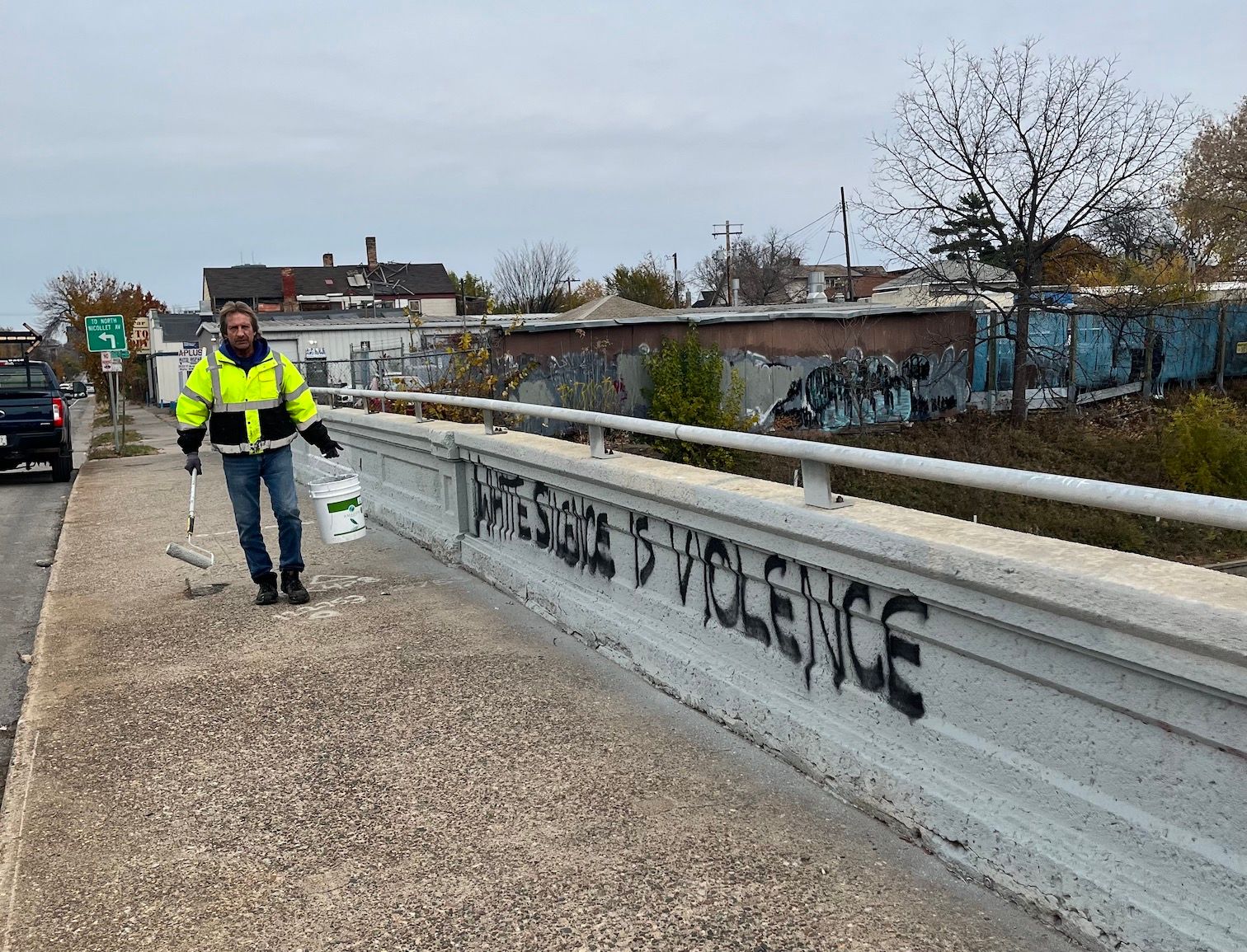 A man with a paint roller approaches a concrete bridge guard. Spray-painted on the bridge is the phrase "White Silence is Violence"