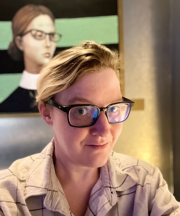 Selfie photo of Deborah Carver, a woman with glasses, in front of a painting of an extremely similar woman with glasses.